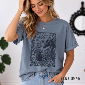 May include: A dusty blue short-sleeve t-shirt featuring a dark navy graphic print of a person reaching toward a dragon surrounded by flowers. The text "One boop, if thou wilt." is printed above the dragon. The shirt has a crew neckline.