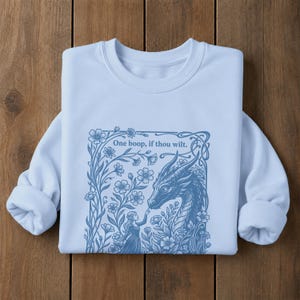 May include: A light blue crewneck sweatshirt featuring a dark blue graphic print of a dragon and a person surrounded by flowers. The text "One boop, if thou wilt." is printed above the dragon. The design is framed by an ornate, rectangular border.