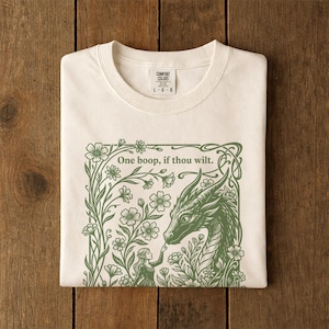 May include: An off-white t-shirt featuring a green graphic print of a dragon and a person surrounded by flowers. The text "One boop, if thou wilt." is printed above the dragon. The shirt has a Comfort Colors brand label at the neck.
