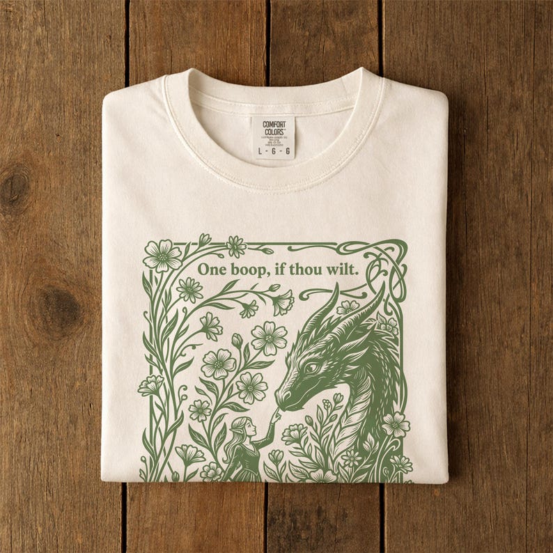May include: An off-white t-shirt featuring a green graphic print of a dragon and a person surrounded by flowers. The text "One boop, if thou wilt." is printed above the dragon. The shirt has a Comfort Colors brand label at the neck.