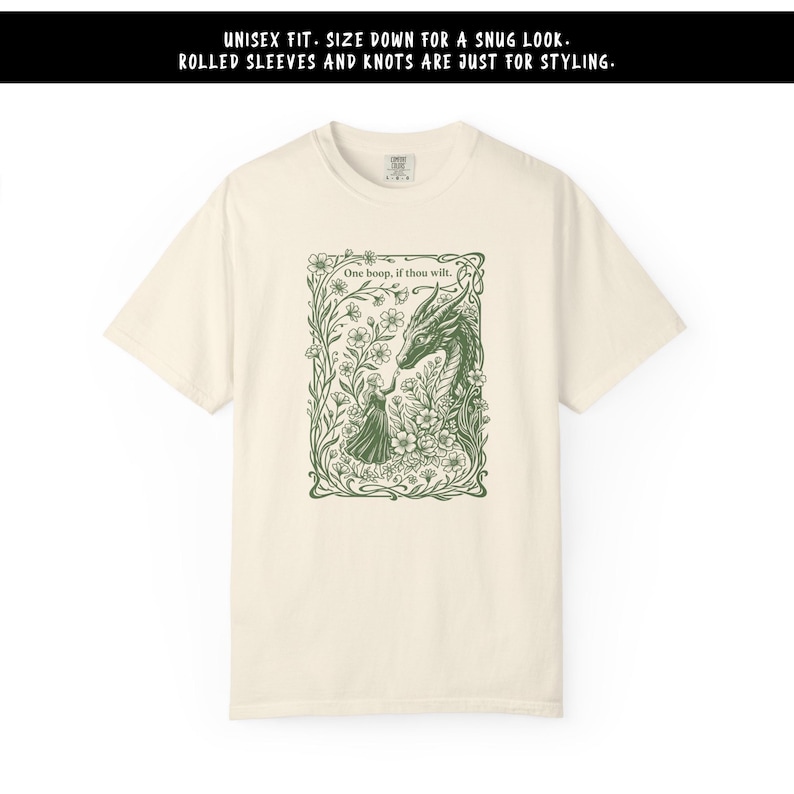 May include: A cream-colored t-shirt featuring a dark green graphic of a person touching a dragon's nose amidst flowers. The text reads: "UNISEX FIT. SIZE DOWN FOR A SNUG LOOK. ROLLED SLEEVES AND KNOTS ARE JUST FOR STYLING." and "One boop, if thou wilt."