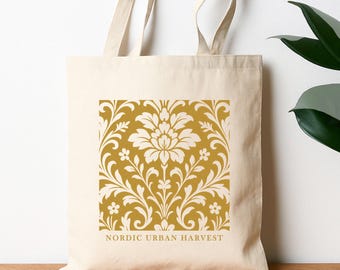 May include: Cream-colored tote bag with a gold floral design. The design features a central flower with surrounding leaves and smaller flowers. The text "NORDIC URBAN HARVEST" is printed below the design.