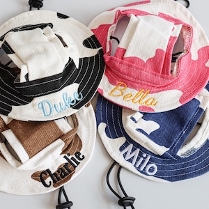 May include: Four personalised pet hats in various colours and patterns. One hat is black and white with "Duke" embroidered in blue. Another is pink and white with "Bella" in yellow. A third is blue and white with "Milo" in white. The last is brown and white with "Charlie" in black.