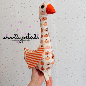 May include: A handmade goose plush toy with a long neck and orange beak. The body is patterned with smiling pumpkins on a cream background. The wings have orange and white stripes, and the legs are orange checkered. The text "woolly petals craftiness and quilts" is visible.
