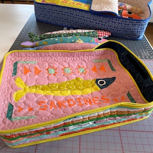 Puede incluir: Un recipiente de tela acolchada rosa con forma de sardina con un dise&ntilde;o de pez amarillo y la palabra "SARDINES" en naranja. El recipiente est&aacute; lleno de peces de tela y se encuentra cerca de un recipiente con estampado azul.