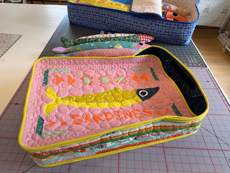 Puede incluir: Un recipiente de tela acolchada rosa con forma de sardina con un dise&ntilde;o de pez amarillo y la palabra "SARDINES" en naranja. El recipiente est&aacute; lleno de peces de tela y se encuentra cerca de un recipiente con estampado azul.