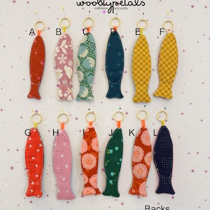 May include: A collection of twelve fish-shaped keychains, each with a unique fabric pattern and color. The keychains are attached to gold-colored rings. The text "woollypetals craftiness and quilts" is at the top.