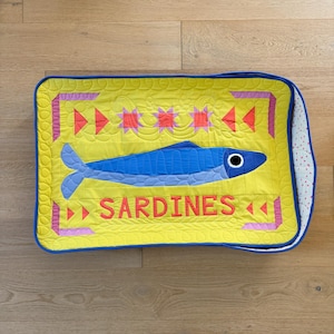 Puede incluir: Un coj&iacute;n rectangular acolchado con fondo amarillo y una ilustraci&oacute;n de sardina azul. La palabra "SARDINES" est&aacute; en letras rojas. El coj&iacute;n tiene un borde azul y un estampado de lunares blancos y rosas en el lateral.