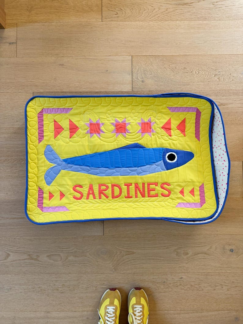 Puede incluir: Un coj&iacute;n rectangular acolchado con fondo amarillo y una ilustraci&oacute;n de sardina azul. La palabra "SARDINES" est&aacute; en letras rojas. El coj&iacute;n tiene un borde azul y un estampado de lunares blancos y rosas en el lateral.