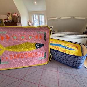 Puede incluir: Dos cajas de tela acolchada con dise&ntilde;os de sardinas. La caja rosa presenta un pez amarillo y la palabra "SARDINES" en naranja. La caja azul tiene un interior amarillo y un dise&ntilde;o de pez azul. Ambas tienen ribetes amarillos.