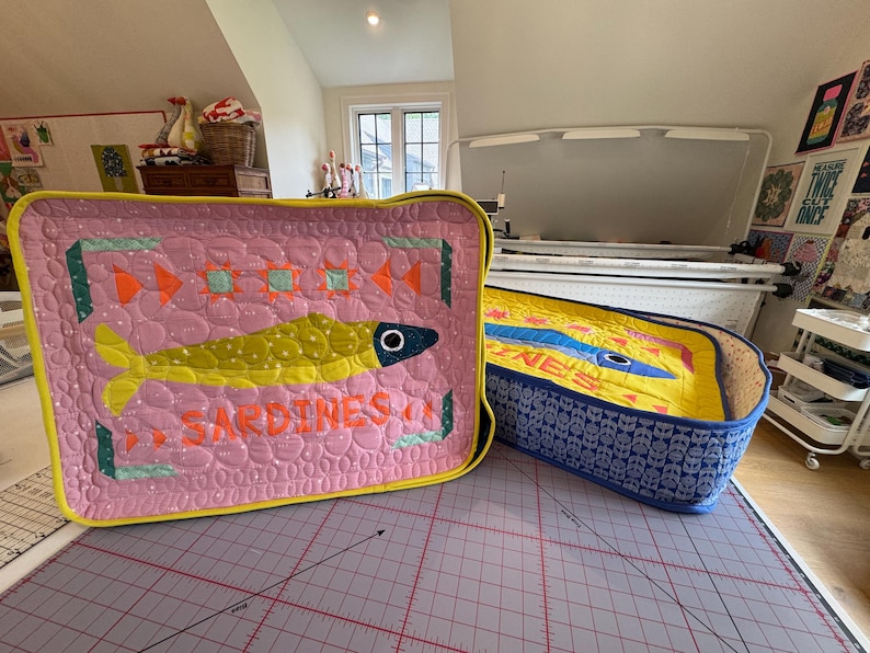 Puede incluir: Dos cajas de tela acolchada con dise&ntilde;os de sardinas. La caja rosa presenta un pez amarillo y la palabra "SARDINES" en naranja. La caja azul tiene un interior amarillo y un dise&ntilde;o de pez azul. Ambas tienen ribetes amarillos.