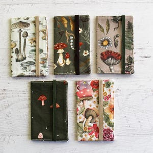 May include: Five fabric-covered notebooks with elastic closures, each featuring a unique botanical design. The covers display mushrooms, flowers, and foliage in various colors, set against backgrounds of white, green, and beige. The notebooks are arranged on a white wooden surface.
