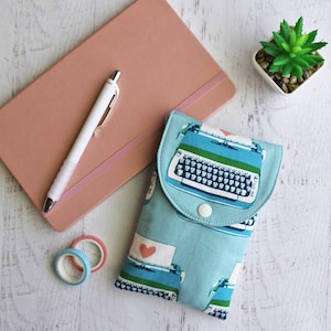 May include: A light blue fabric case with a typewriter design, a pink notebook, a white pen, a small potted succulent, and decorative tape rolls. The case has a button closure and a heart design.