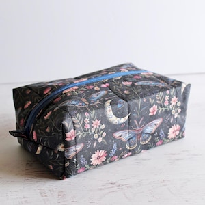 May include: A rectangular cosmetic bag with a dark background featuring a floral and moth pattern. The bag has a blue zipper and is likely made of fabric. The design includes pink flowers, moths, and a crescent moon.