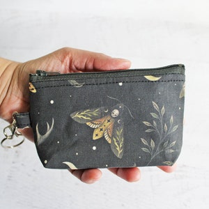 May include: A small, dark gray zippered pouch with a black and white moth design and a silver key chain. The pouch has a black and white starry night background with a floral pattern.