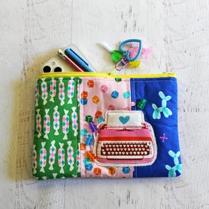 May include: A colorful zippered pouch with a patchwork design. The pouch features a green candy pattern, pink cubes, and blue balloon dog designs. A pink typewriter patch with a blue heart is on the front. A phone and pen are visible inside.
