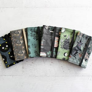 May include: A collection of fabric-covered journals with elastic closures, featuring various patterns. Designs include black cats, bats, botanical prints, and a witchy illustration. The journals are arranged in a semi-circle on a white wooden surface.