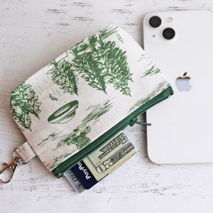 May include: A white zippered pouch with a green and white floral print. The pouch has a silver key ring attached and is open to reveal a twenty dollar bill and a credit card.