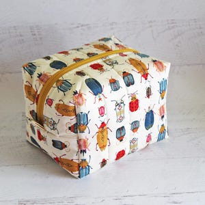 May include: A cream-colored, rectangular zippered pouch with a repeating pattern of colorful beetles. The bugs are in shades of red, blue, yellow, and brown. The zipper is yellow.