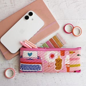 May include: A pink quilted pencil case featuring embroidered designs of a typewriter, a fried egg, a gummy bear, and an ice cream cone. The case is accompanied by a white smartphone, a pink notebook, three pens, two rolls of washi tape, and sticky notes.