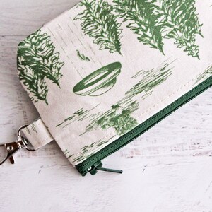 Puede incluir: Una peque&ntilde;a bolsa de cremallera de color crema con una cremallera verde y un estampado floral verde. La bolsa tiene un clip de metal plateado.