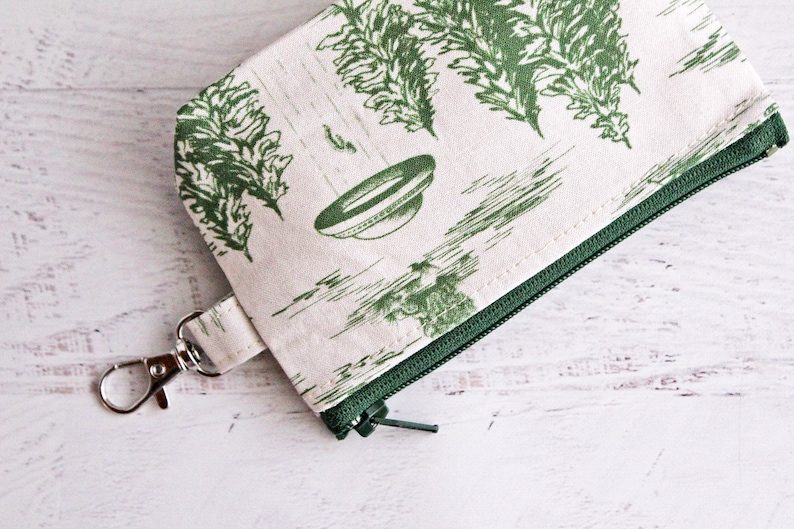 Puede incluir: Una peque&ntilde;a bolsa de cremallera de color crema con una cremallera verde y un estampado floral verde. La bolsa tiene un clip de metal plateado.