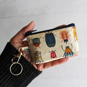 May include: A small beige zippered pouch with a key ring. The pouch is decorated with a colorful pattern of various insects.