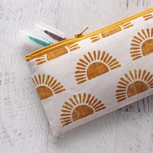 May include: A white canvas zippered pouch with a yellow zipper and a repeating pattern of yellow suns. The pouch is lying on a white wooden surface.
