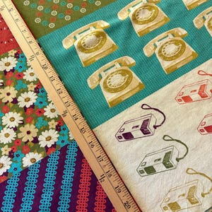May include: A multi-patterned fabric featuring sections of green floral print, teal with yellow rotary telephones, white with purple and green cassette players, and purple with teal wavy stripes, measured by a wooden ruler up to 22 inches.