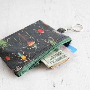 May include: A small black zippered pouch with a colorful insect print. The pouch is open and contains cash and a credit card. The pouch has a key ring attached.