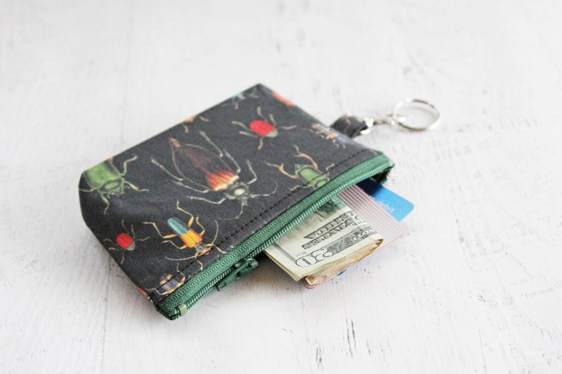 May include: A small black zippered pouch with a colorful insect print. The pouch is open and contains cash and a credit card. The pouch has a key ring attached.