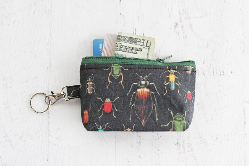 May include: A small black zippered pouch with a key ring. The pouch has a print of colorful beetles on a black background. The pouch is partially open, revealing a twenty dollar bill and a debit card.