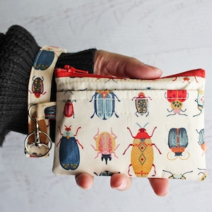 May include: A small white zippered pouch with a red zipper and a colorful beetle print. The pouch is attached to a wristlet with a silver key ring.