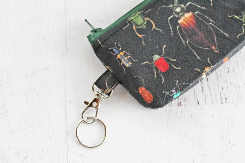 May include: A black fabric zippered pouch with a green zipper pull and a silver key ring. The pouch is decorated with a repeating pattern of colorful insects.