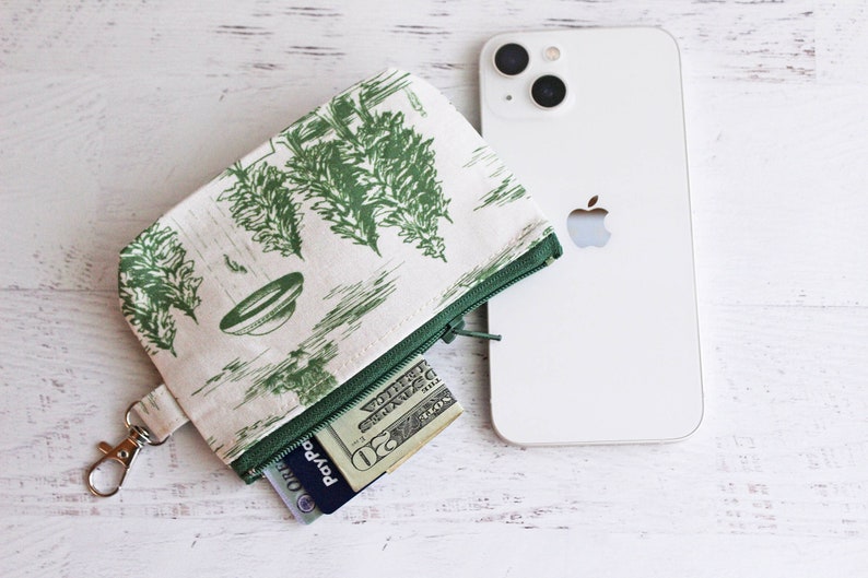 Puede incluir: Una bolsa blanca con cremallera con un estampado floral verde y blanco. La bolsa tiene un llavero plateado y est&aacute; abierta para revelar un billete de veinte euros y una tarjeta de cr&eacute;dito.