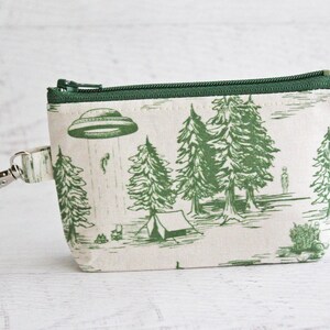 Puede incluir: Una bolsa de cremallera verde y blanca con un estampado de un OVNI, &aacute;rboles y una tienda de campa&ntilde;a. La bolsa tiene un llavero plateado.