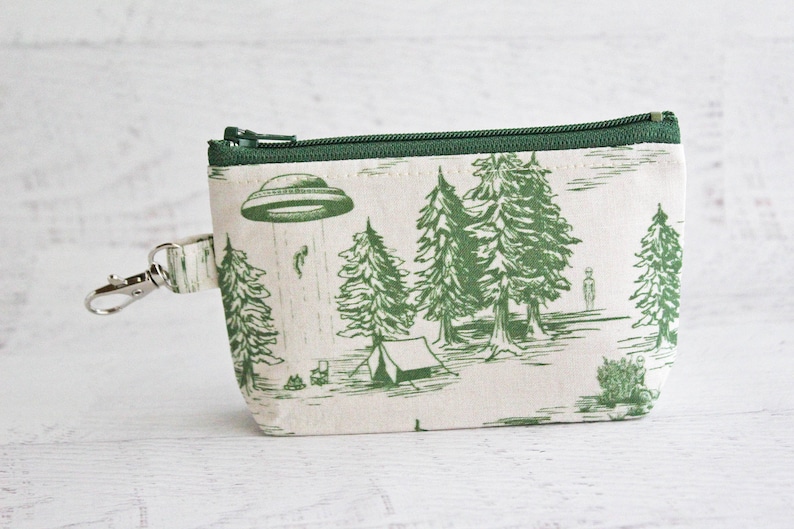 Puede incluir: Una bolsa de cremallera verde y blanca con un estampado de un OVNI, &aacute;rboles y una tienda de campa&ntilde;a. La bolsa tiene un llavero plateado.