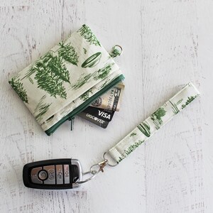 May include: A small green and white fabric pouch with a zipper closure and a matching wrist strap. The pouch is open and a credit card is visible inside. A key fob is also pictured.