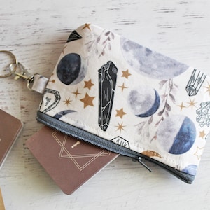 May include: A white zippered pouch with a moon, stars, and crystal pattern. The pouch has a silver key ring attached to it. The pouch is lying on a white wooden surface.