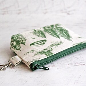 Puede incluir: Una peque&ntilde;a bolsa de cremallera blanca con estampado floral verde y un llavero plateado. La bolsa tiene una cremallera verde y un fondo blanco.