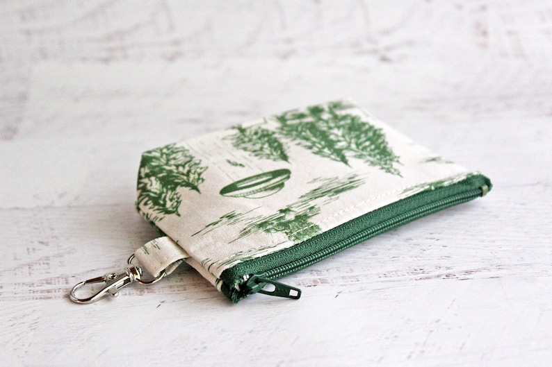 Puede incluir: Una peque&ntilde;a bolsa de cremallera blanca con estampado floral verde y un llavero plateado. La bolsa tiene una cremallera verde y un fondo blanco.