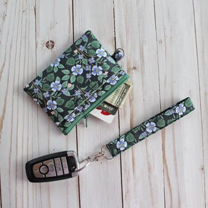 May include: A green and blue floral fabric wallet with a zipper closure and a matching wrist strap. The wallet has a credit card and cash inside.