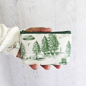 Puede incluir: Una bolsa blanca con cremallera y ribete verde. La bolsa tiene un estampado verde y blanco de un OVNI, &aacute;rboles y una tienda de campa&ntilde;a. La bolsa tiene un tirador de cremallera plateado con un anillo.