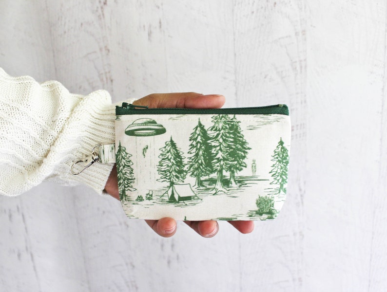 Puede incluir: Una bolsa blanca con cremallera y ribete verde. La bolsa tiene un estampado verde y blanco de un OVNI, &aacute;rboles y una tienda de campa&ntilde;a. La bolsa tiene un tirador de cremallera plateado con un anillo.
