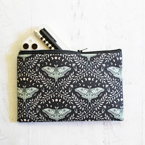 May include: Black zippered pouch with a repeating pattern of moths, moons, and foliage in white and pale green. A smartphone and pen are partially visible inside the open pouch. The pouch is rectangular and lies on a white surface.
