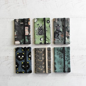 May include: Six fabric card holders with elastic closures. The holders feature various patterns, including black cats, bats, and botanical designs. Colors include black, green, and gray. The holders are rectangular and appear to be handmade.