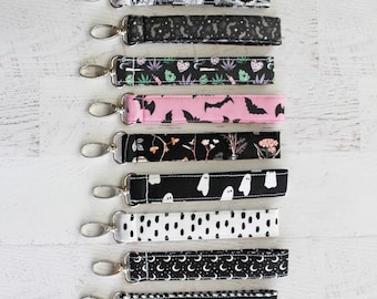 May include: A set of eight black and white keychains with different spooky patterns, including bats, ghosts, moons, and stars. The keychains are made of fabric and have silver key rings.