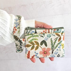 May include: A small floral print zippered pouch with a key chain attached. The pouch is made of a white fabric with a green border and features a variety of colorful flowers and leaves.