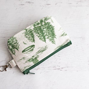 Puede incluir: Una peque&ntilde;a bolsa de cremallera blanca con una cremallera verde y un estampado floral verde. La bolsa tiene un llavero plateado.