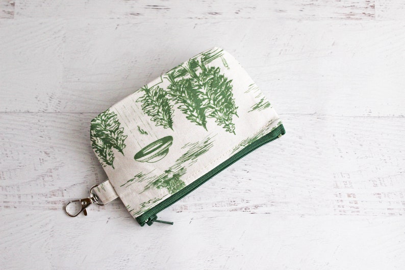 Puede incluir: Una peque&ntilde;a bolsa de cremallera blanca con una cremallera verde y un estampado floral verde. La bolsa tiene un llavero plateado.